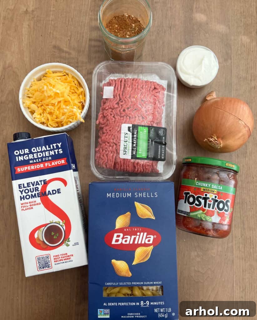 Quick One-Pot Cheesy Taco Pasta 3 A flat lay image showing the key ingredients for taco pasta: ground beef, shell pasta, beef broth, cheese, taco seasoning, onion, sour cream, and salsa.