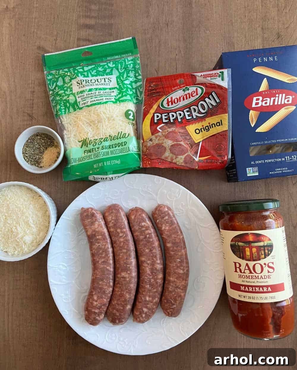 A colorful array of ingredients for pizza pasta bake, including Italian sausage, marinara sauce, uncooked penne pasta, pepperoni, various spices, and shredded cheese.