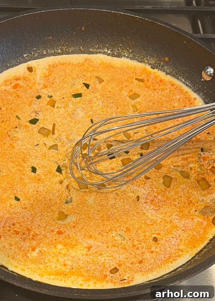 Heavy cream being whisked with sautéed spices in a cast iron skillet, gently heating to combine flavors before cheese is added.