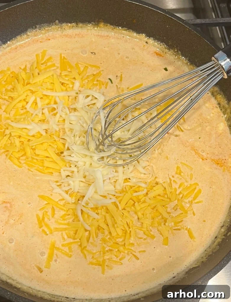 Shredded pepper jack and cheddar cheese melting into the creamy sauce in a cast iron skillet, being whisked until smooth to create homemade queso.