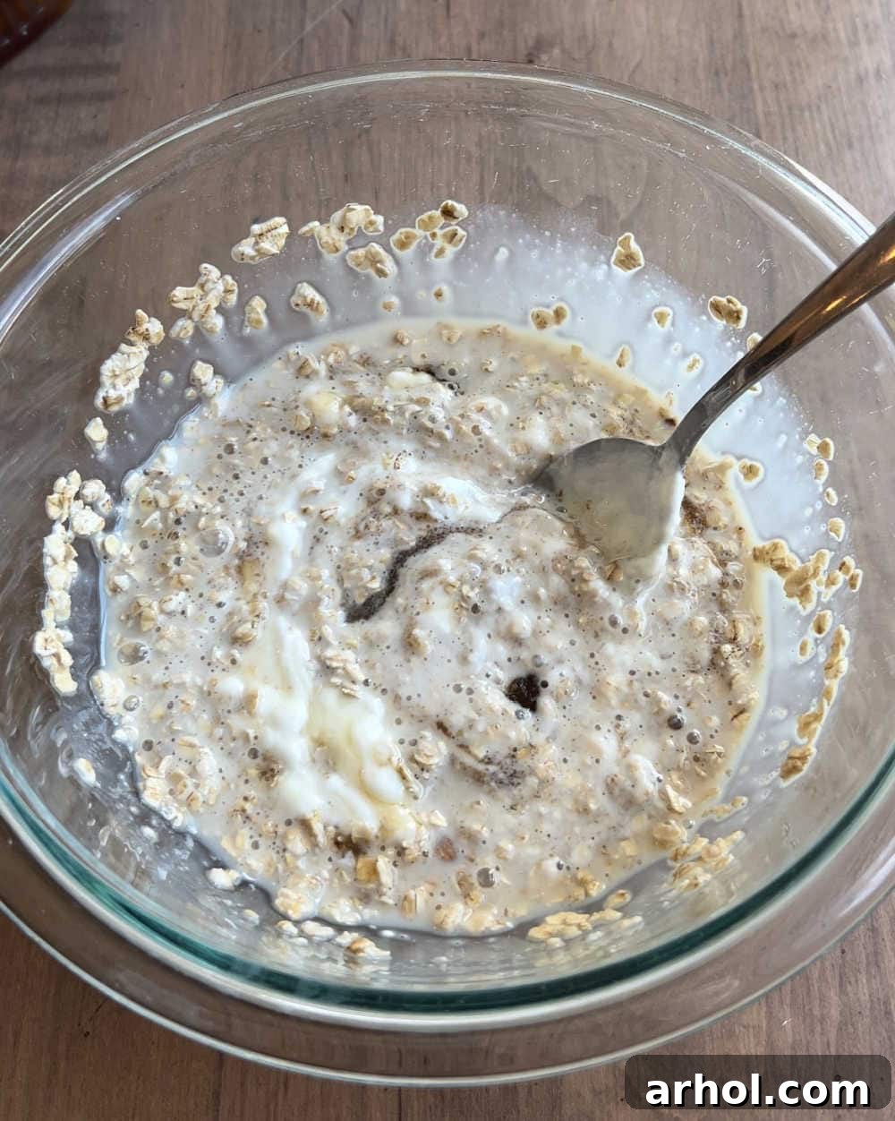 A hand stirring a mixture of oats, milk, yogurt, brown sugar, and maple syrup in a clear glass mixing bowl.
