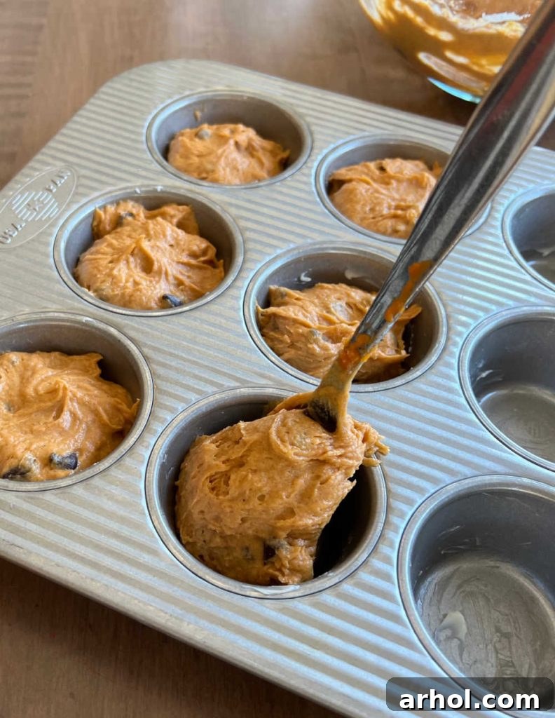 Hands spooning the thick pumpkin muffin batter into a lined muffin tin, ready for baking.