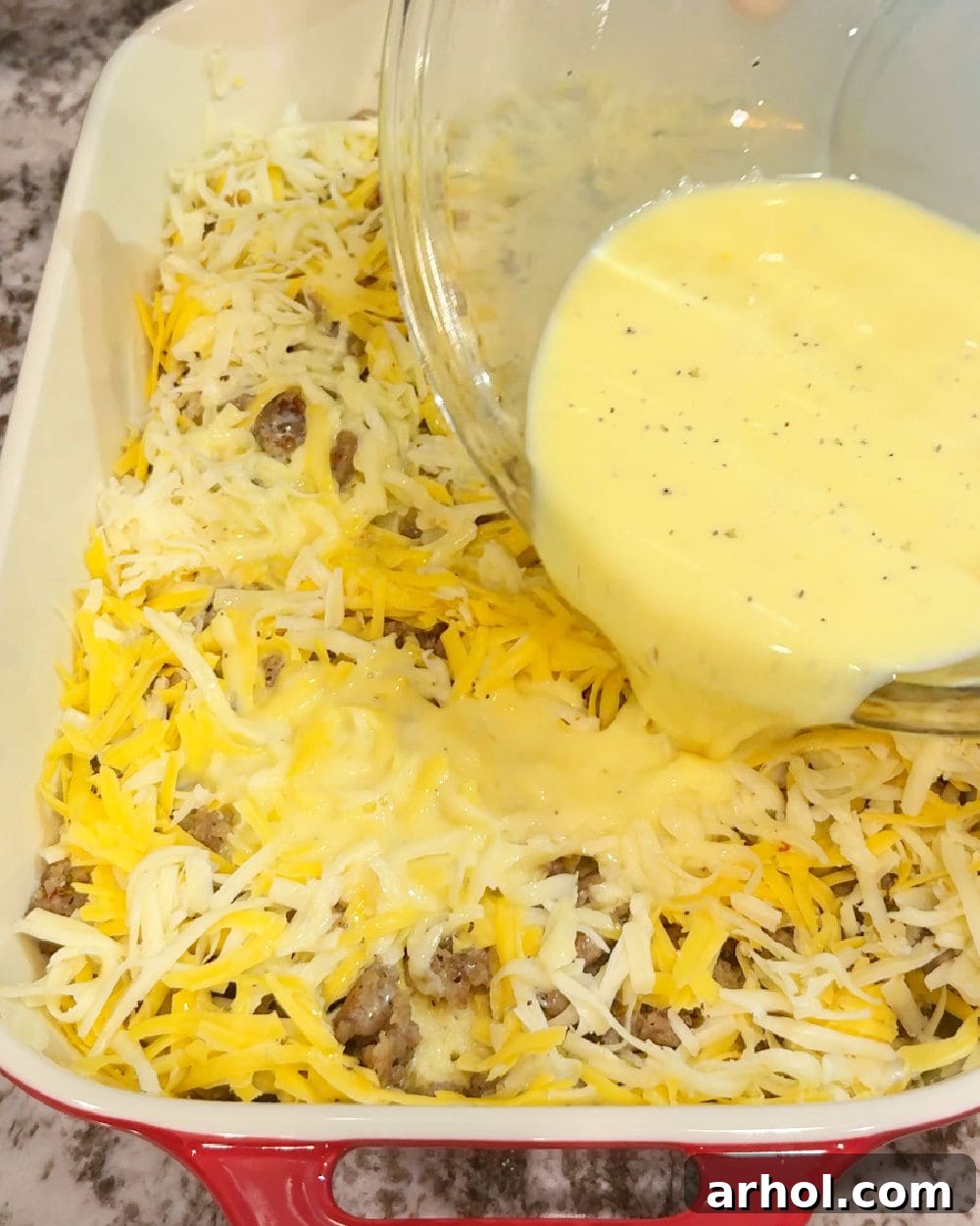 Pouring the whisked egg mixture evenly over the bread, sausage, and cheese layers in the baking dish.