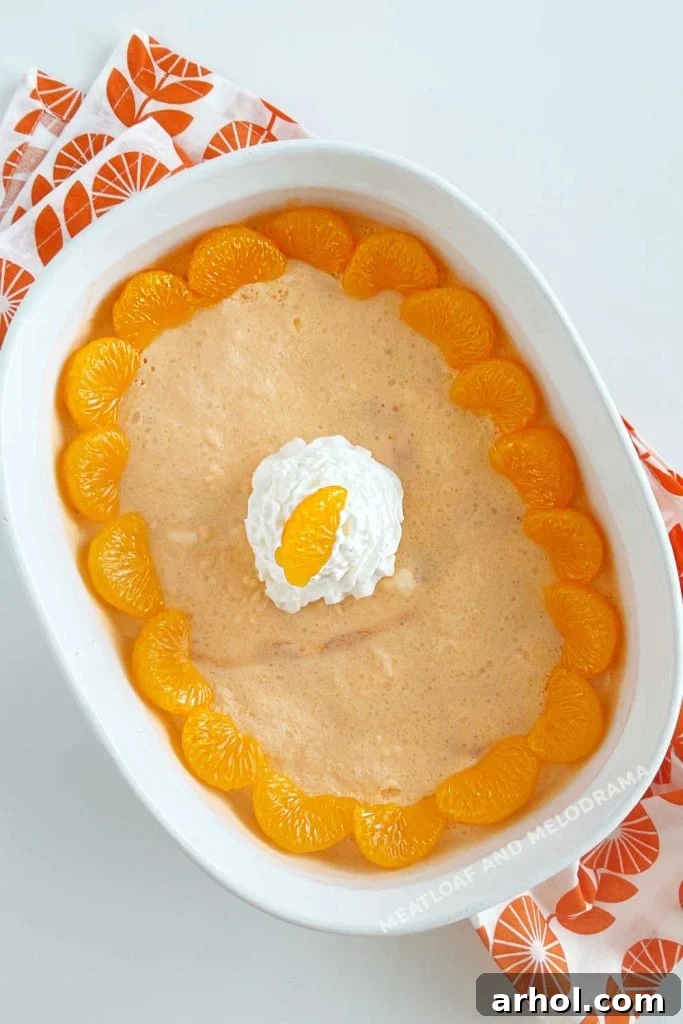 A serving dish filled with creamy orange jello salad, topped with a dollop of whipped cream and a mandarin orange slice, ready for serving.
