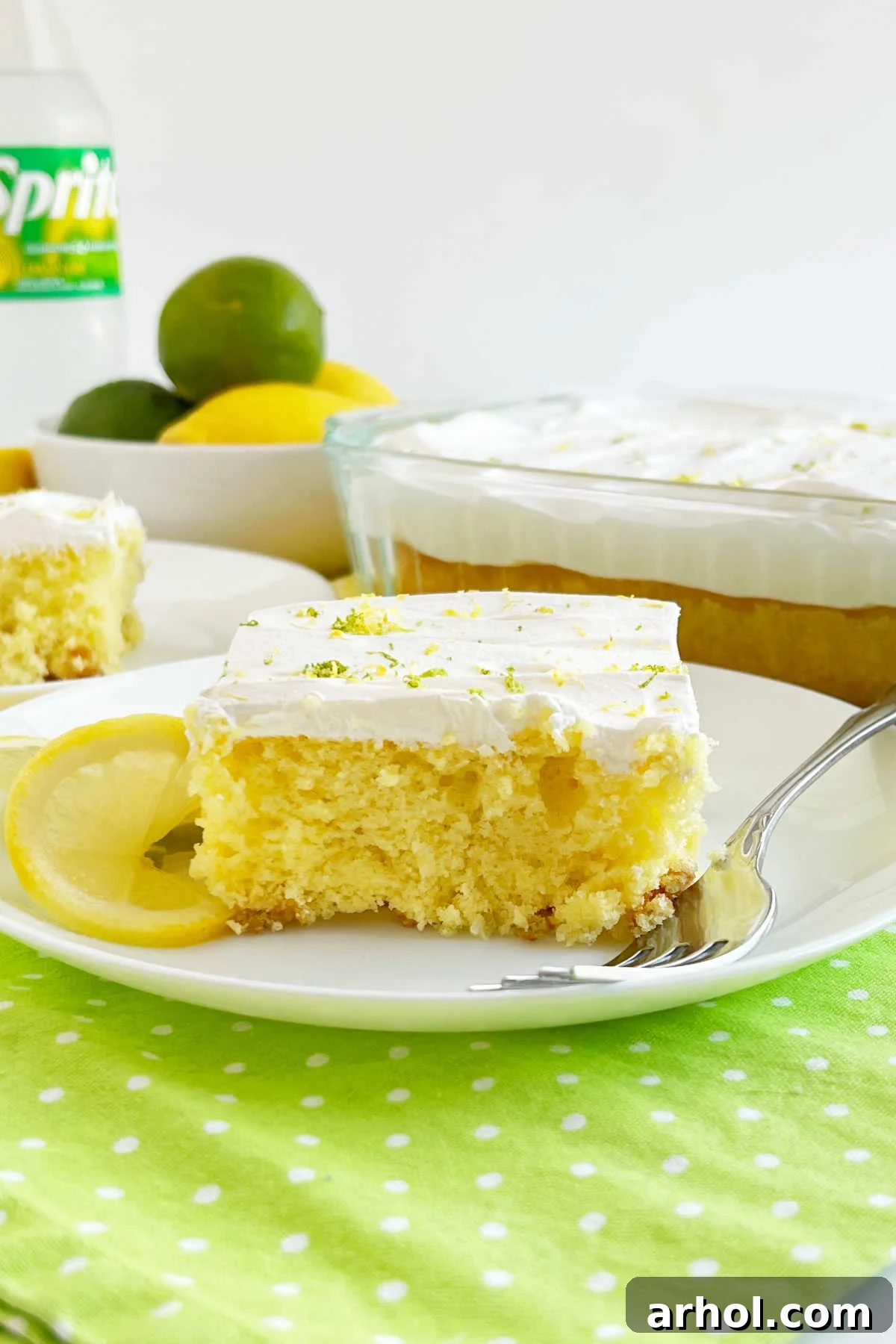 A vibrant slice of Lemon Lime Soda Cake, generously frosted with cool whipped topping and garnished with fresh lemon and lime zest, served on a pristine white plate.