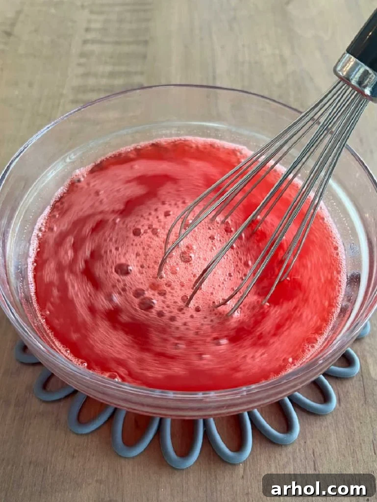 Strawberry Jello powder being mixed with hot and cold water in a small bowl, creating the vibrant red gelatin liquid for the poke cake.