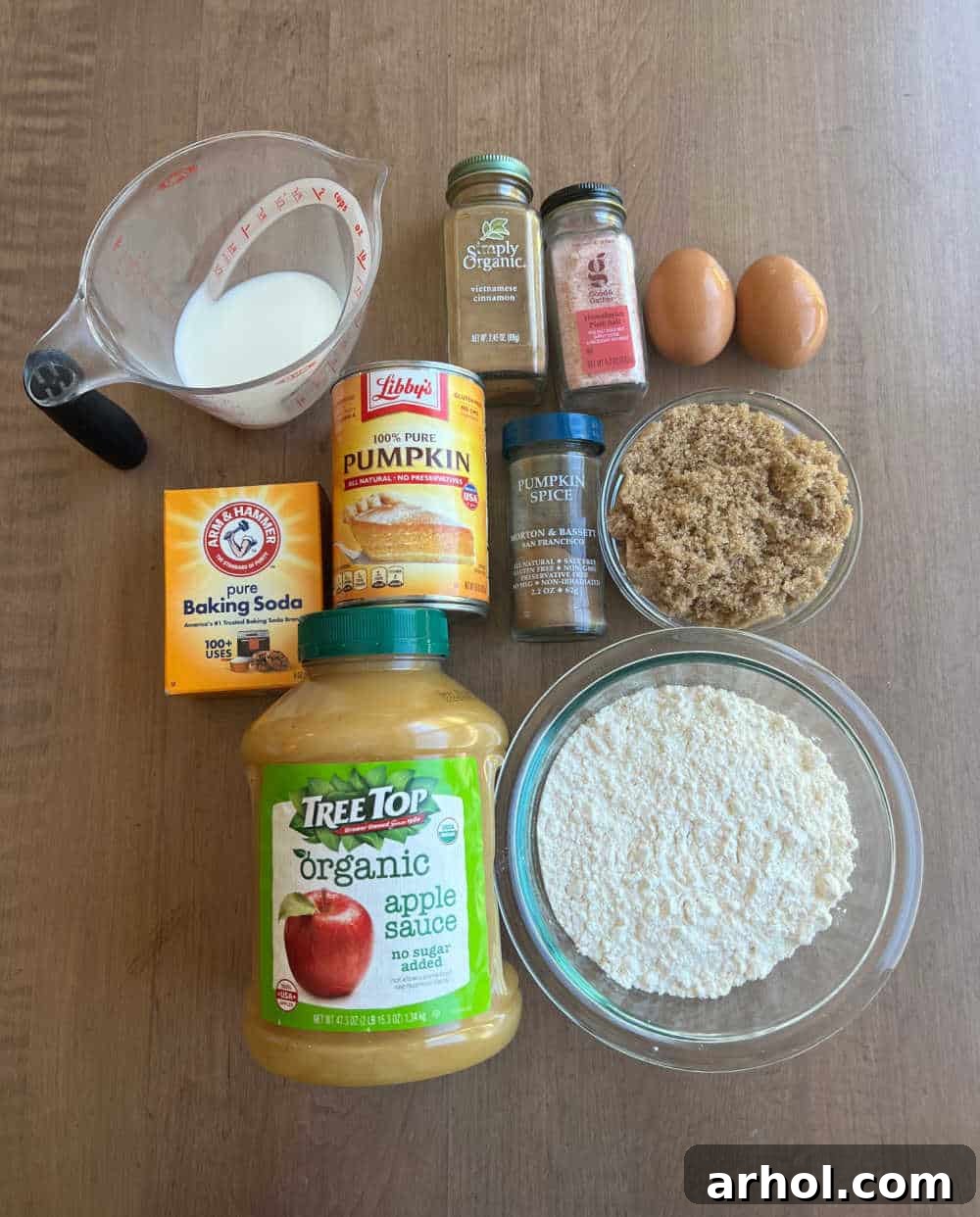 Array of fresh ingredients including canned pumpkin puree, unsweetened applesauce, all-purpose flour, brown sugar, baking soda, milk, eggs, and various spices arranged for making homemade pumpkin muffins.