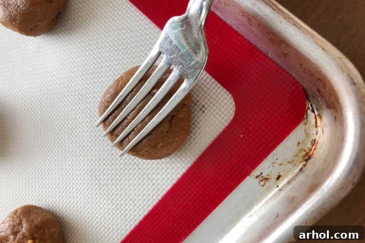 The Easiest 4 Ingredient Peanut Butter Cookies 4 A fork pressing down on peanut butter cookie dough balls, creating the iconic criss-cross pattern on a baking sheet.