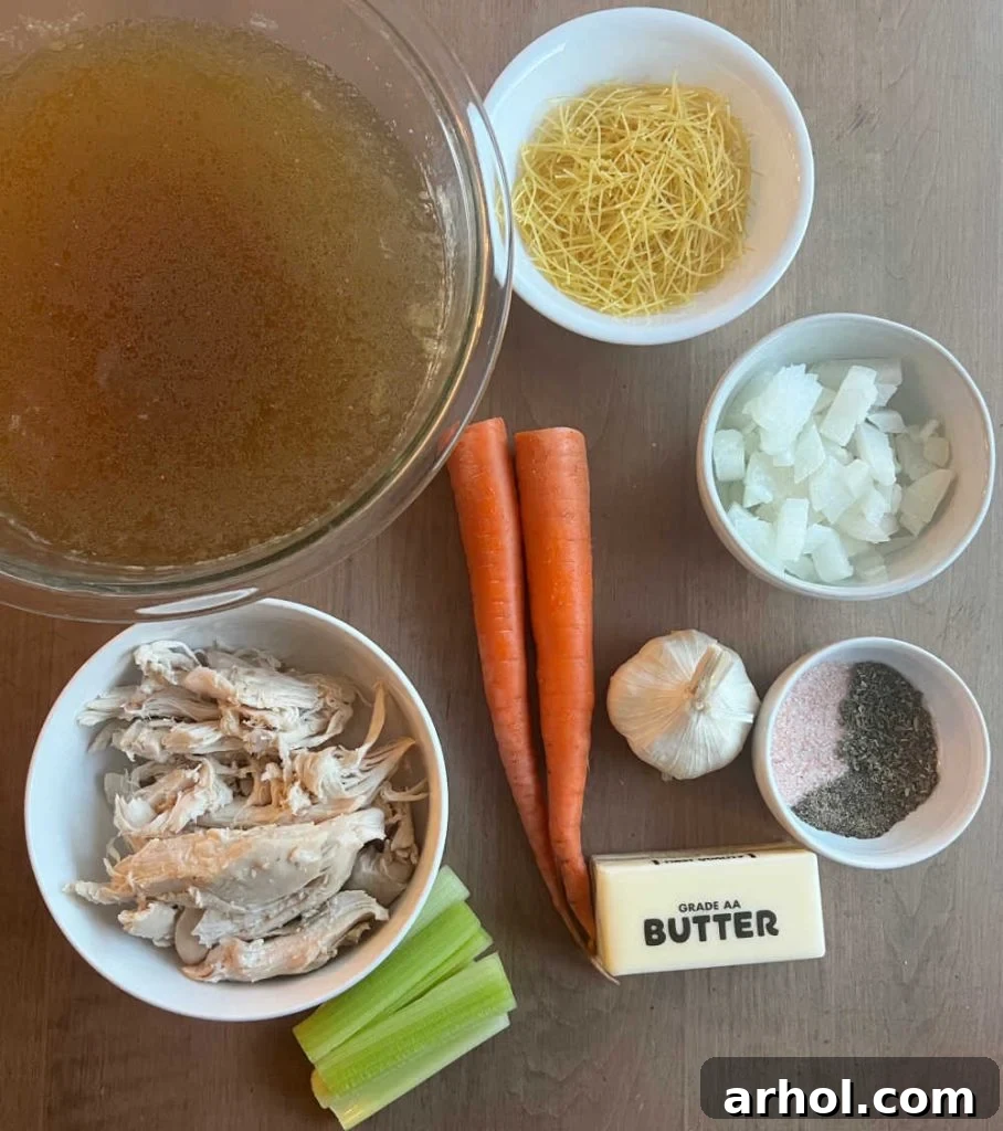 A collection of fresh and prepared ingredients for homemade chicken noodle soup, including homemade chicken stock, shredded rotisserie chicken meat, various noodles, diced onion, sliced carrots, minced garlic, a pat of butter, and a selection of seasonings, all arranged on a rustic surface.