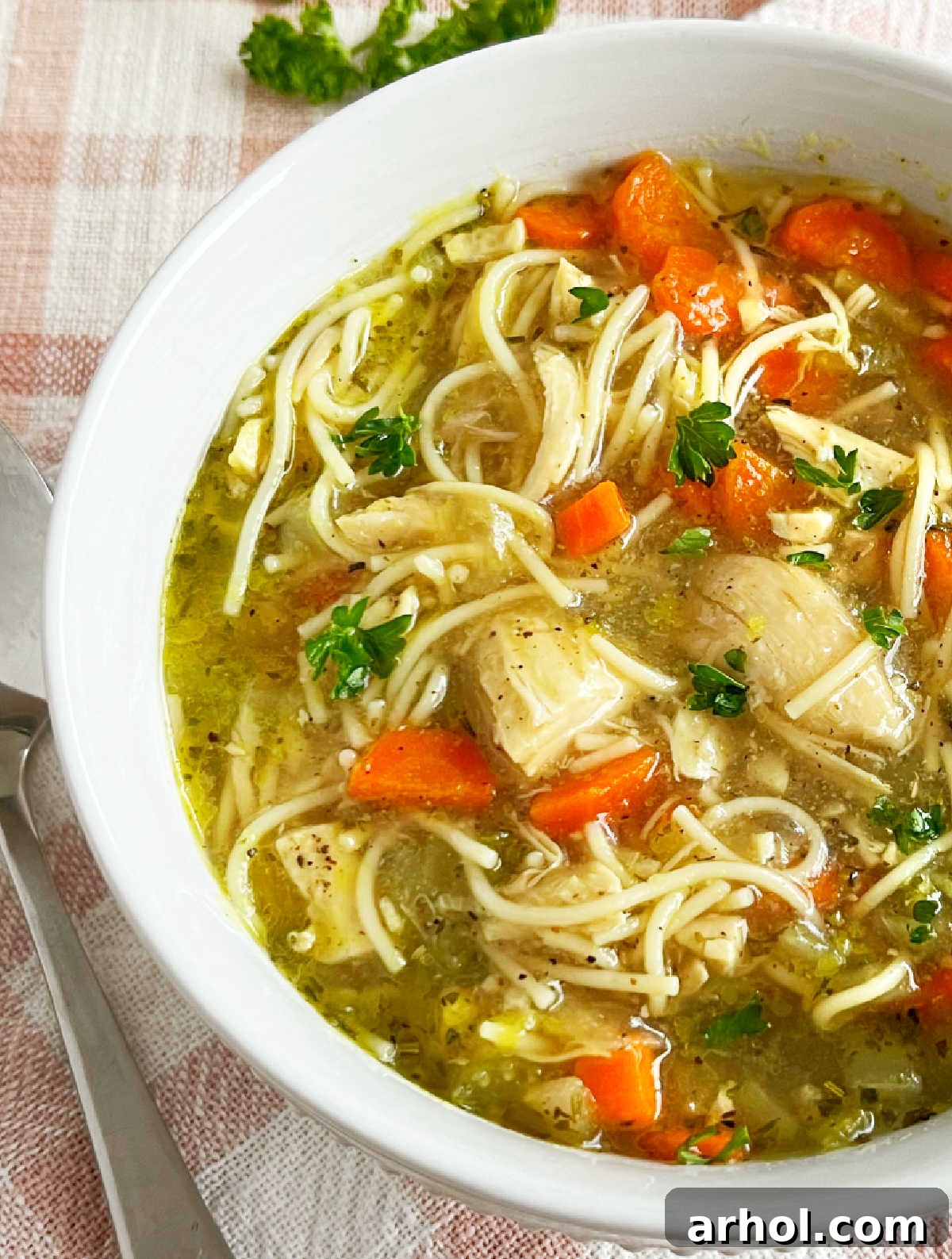 A close-up of a rustic white bowl filled with steaming homemade chicken noodle soup, generously featuring visible carrots, tender shredded chicken, and a vibrant fresh parsley garnish, ready to be enjoyed.