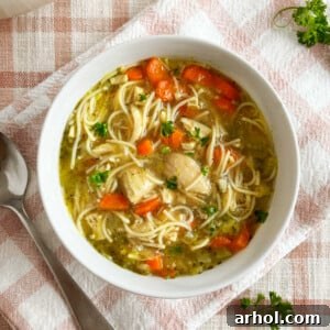 A close-up of a rustic white bowl filled with steaming homemade chicken noodle soup, generously featuring visible carrots, tender shredded chicken, and a vibrant fresh parsley garnish, ready to be enjoyed as a comforting meal.