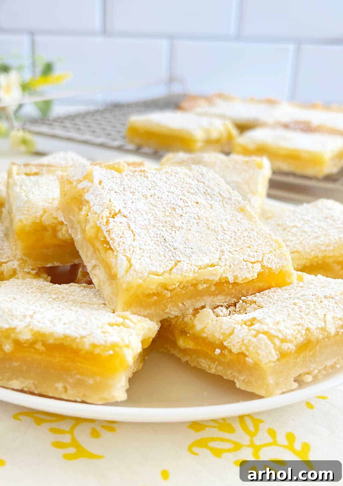 A plate of freshly baked homemade lemon bars, showcasing their golden crust and bright yellow filling, garnished with powdered sugar, resting on a kitchen counter.