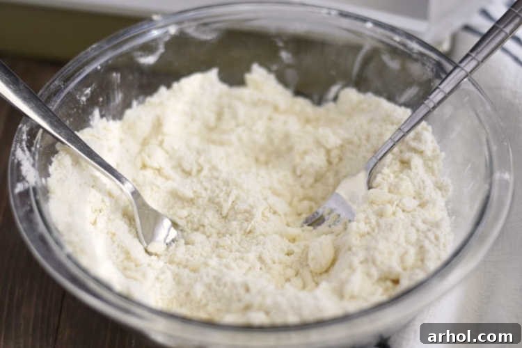 Close-up shot of cold butter being cut into flour and powdered sugar mixture with two forks for a shortbread crust, showing a crumbly texture.