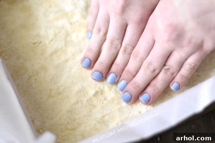 Hands pressing a shortbread crust mixture firmly and evenly into a baking pan lined with parchment paper.