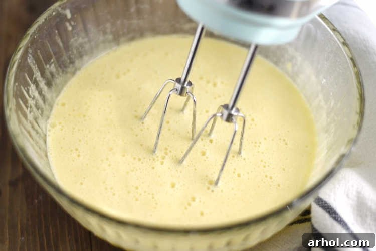 A hand mixer blending the lemon curd filling ingredients – eggs, sugar, flour, lemon juice, and zest – in a large mixing bowl, creating a pale yellow, smooth mixture.