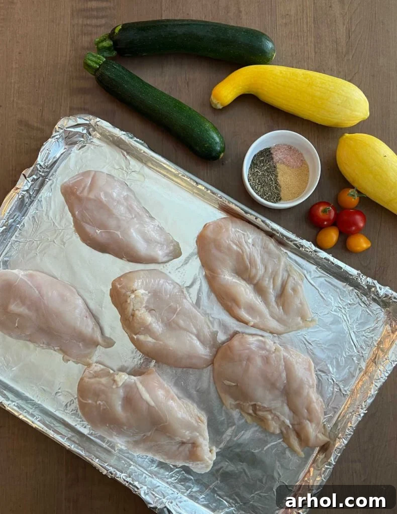 Raw ingredients for baked chicken and zucchini: boneless skinless chicken breasts, sliced zucchini, yellow squash, grape tomatoes, and a mix of herbs and spices.