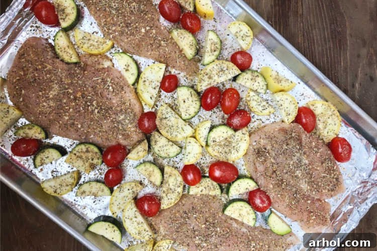 Chicken breasts, sliced zucchini, yellow squash, and grape tomatoes are arranged and seasoned on a baking sheet, prepped for oven roasting.