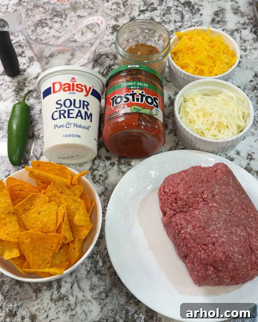 Individual ingredients laid out for Doritos casserole: ground beef, a bag of Doritos, shredded cheese, salsa, taco seasoning, sour cream, and a jalapeño.