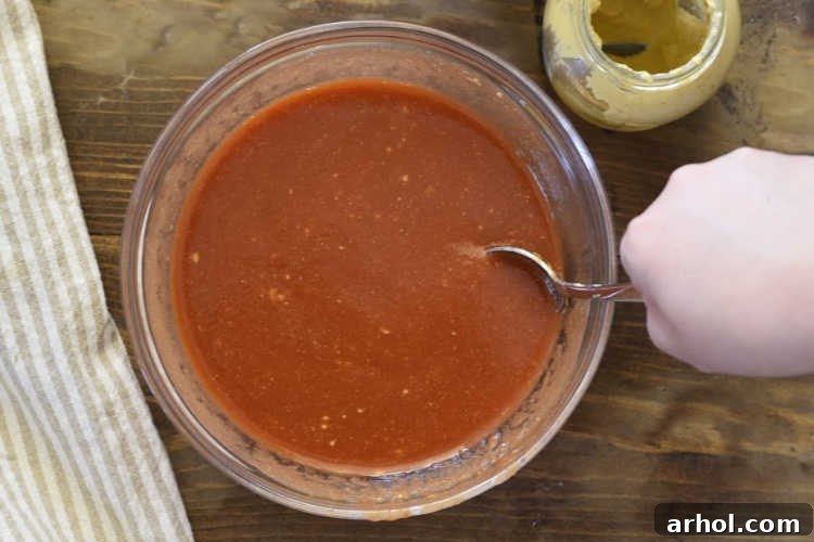 Close-up of a small glass bowl filled with a rich, reddish-brown mixture of tomato sauce, brown sugar, mustard, and vinegar, ready to be whisked into a tangy glaze for meatloaf.