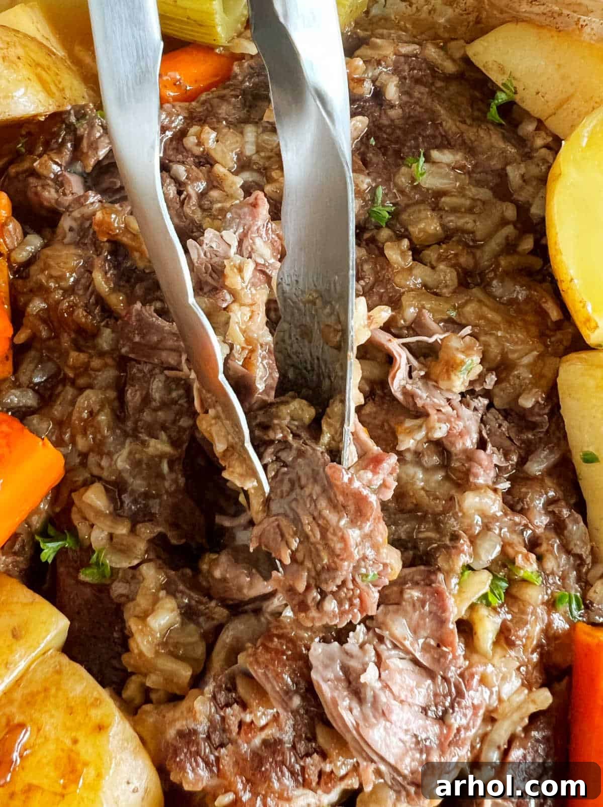 Kitchen tongs gently holding a piece of baked chuck roast, showcasing its tenderness and readiness to be shredded.