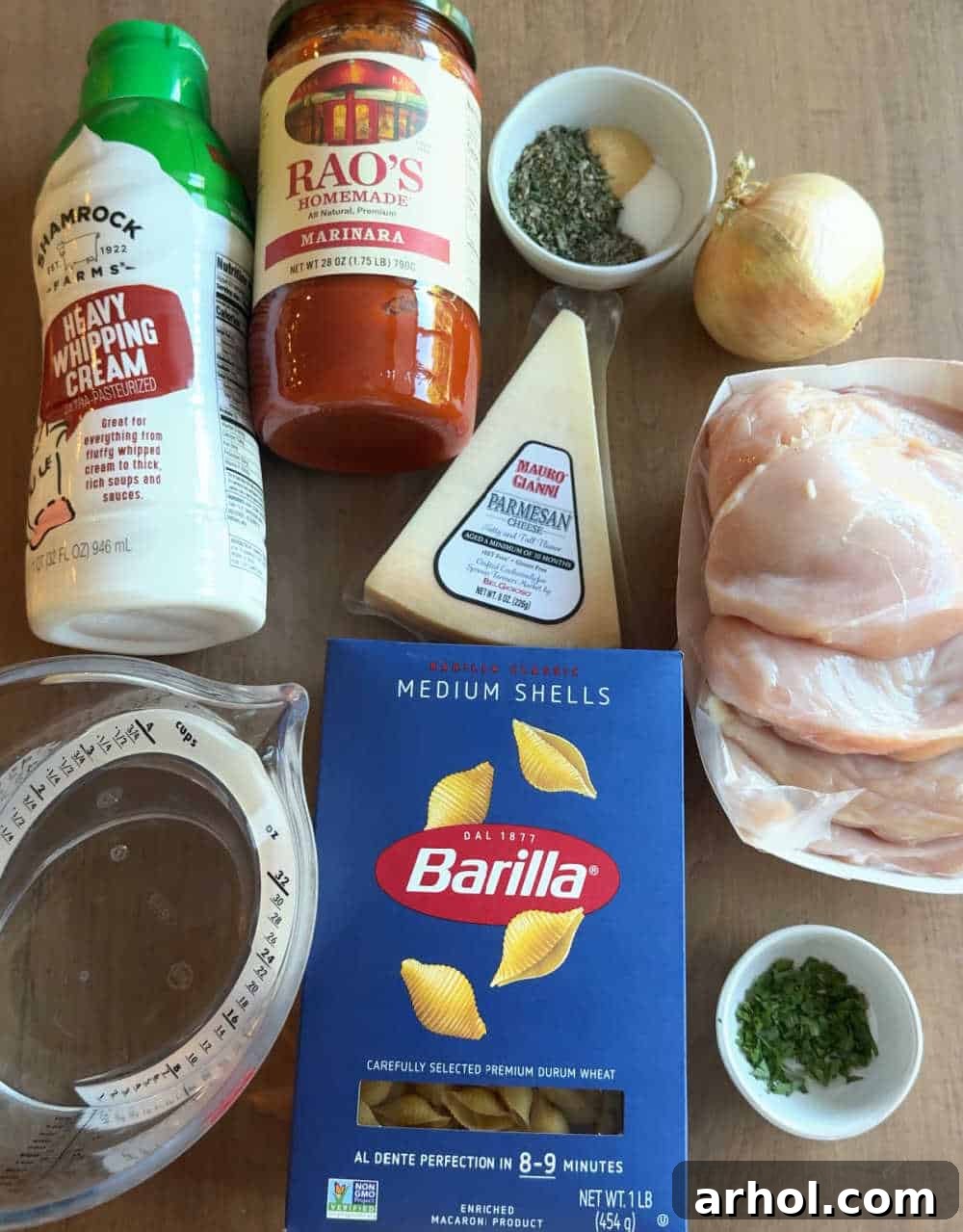 A flat lay photograph showcasing the key ingredients for creamy chicken pasta: raw chicken breast, medium shell pasta, a jar of marinara sauce, a container of heavy cream, a glass of water, and various seasonings.