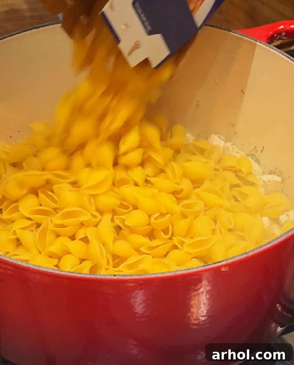 Medium shell pasta being poured into a Dutch oven with cooked chicken and onions.