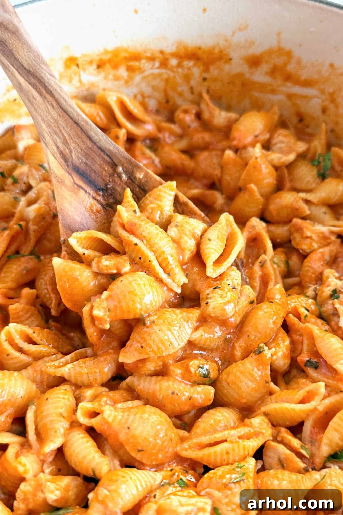 A wooden spoon lifting a spoonful of creamy chicken pasta and shell pasta from a red Dutch oven, showcasing the rich sauce.