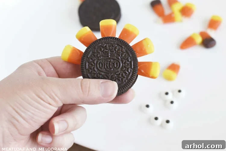 A hand gently pressing a piece of candy corn into the cream filling of an Oreo cookie, forming the turkey's tail feathers.