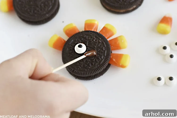 A toothpick applying a small dot of melted chocolate to an Oreo cookie, preparing to attach a candy eye for the turkey's face.