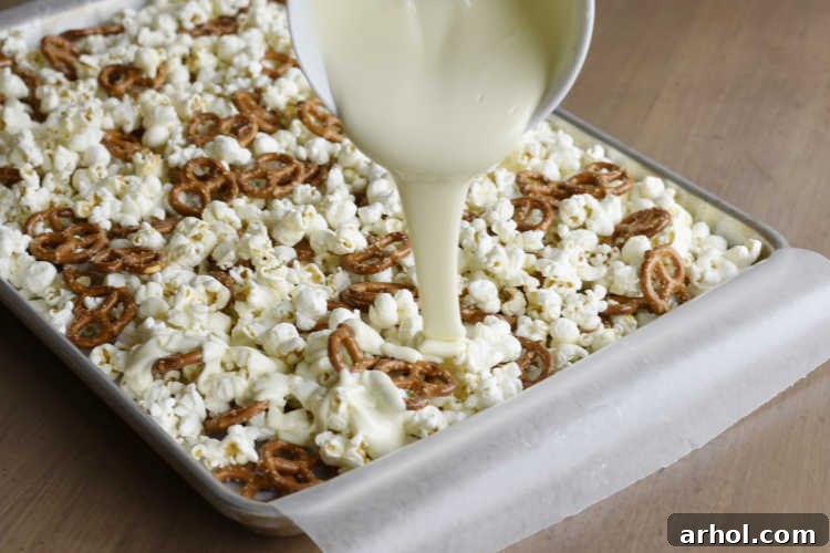 A visual guide showing melted white chocolate being generously poured over a bed of crisp pretzels and fluffy popcorn spread on a baking sheet, ready for mixing.