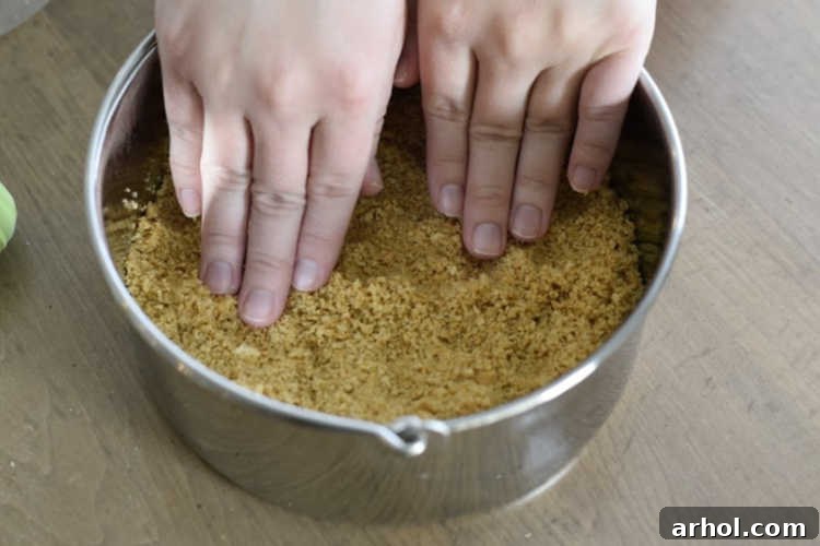 Hands pressing graham cracker crust into a 7-inch springform pan, creating a firm base for the cheesecake.