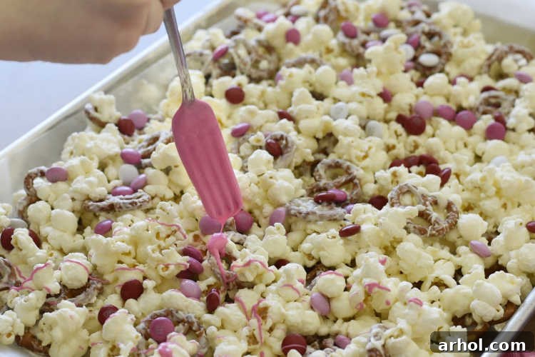 Sweetheart Snack Medley 5 A hand drizzling vibrant pink candy coating over a baking sheet full of popcorn and pretzel snack mix.