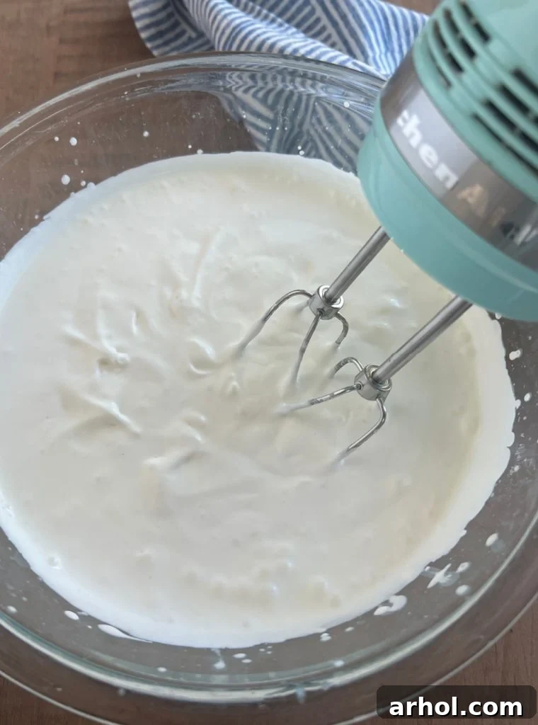 Cold heavy whipping cream being beaten in a large mixing bowl with an electric mixer until stiff peaks form.