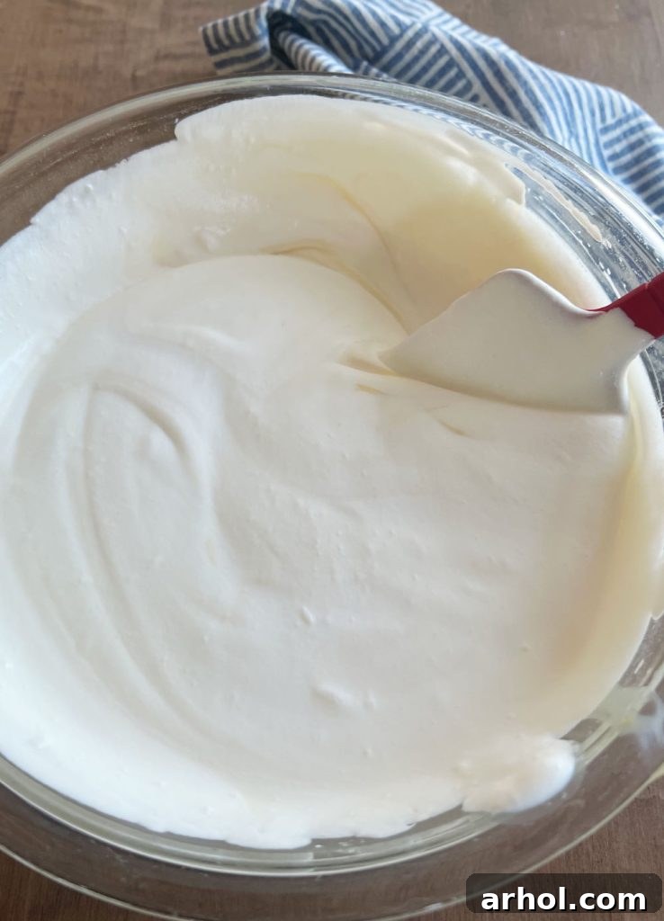Whipped cream being gently folded into sweetened condensed milk in a large mixing bowl with a spatula.