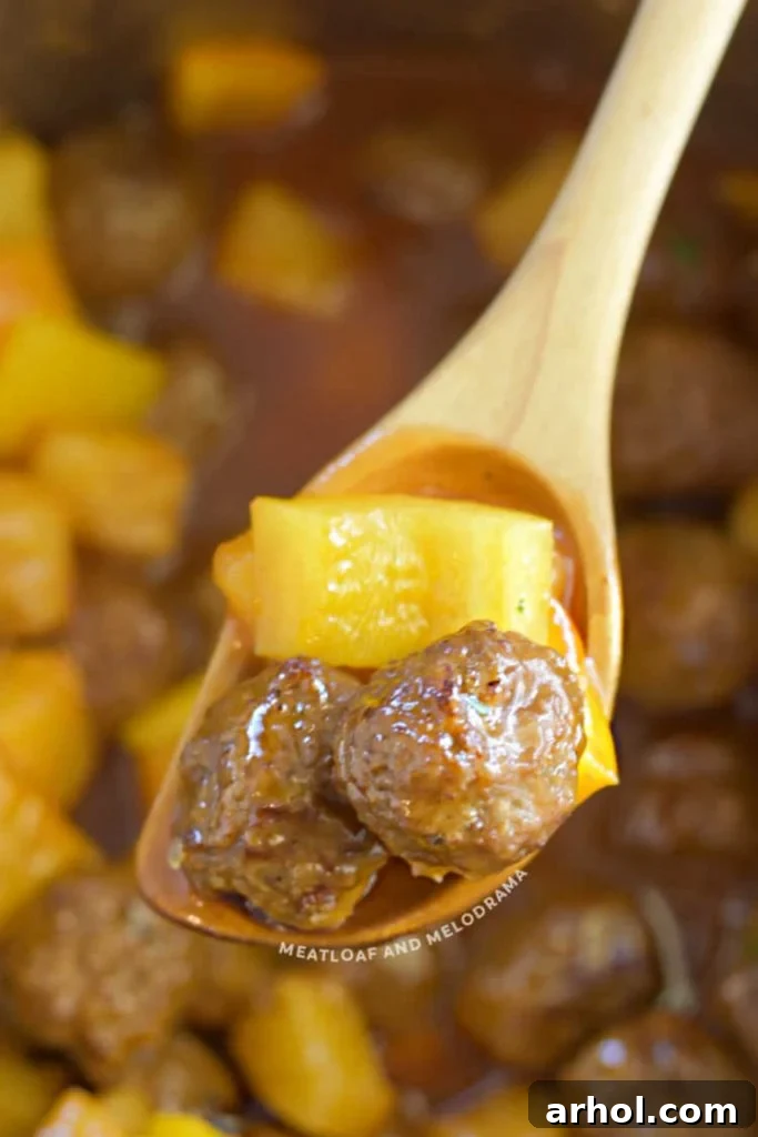 A wooden spoon holding sweet and sour meatballs with pineapple chunks, ready to serve.