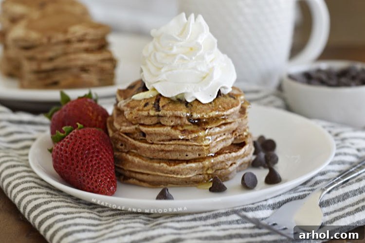 A beautifully plated stack of chocolate chip pancakes topped with whipped cream and fresh strawberries on a white plate, ready to be devoured.