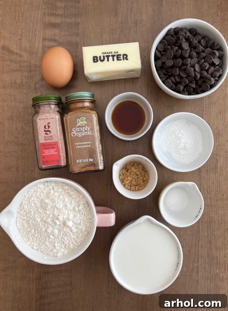 Various ingredients for chocolate chip pancakes laid out on a rustic surface: flour, butter, chocolate chips, baking powder, milk, sugar, brown sugar, vanilla extract, egg, salt, and cinnamon.