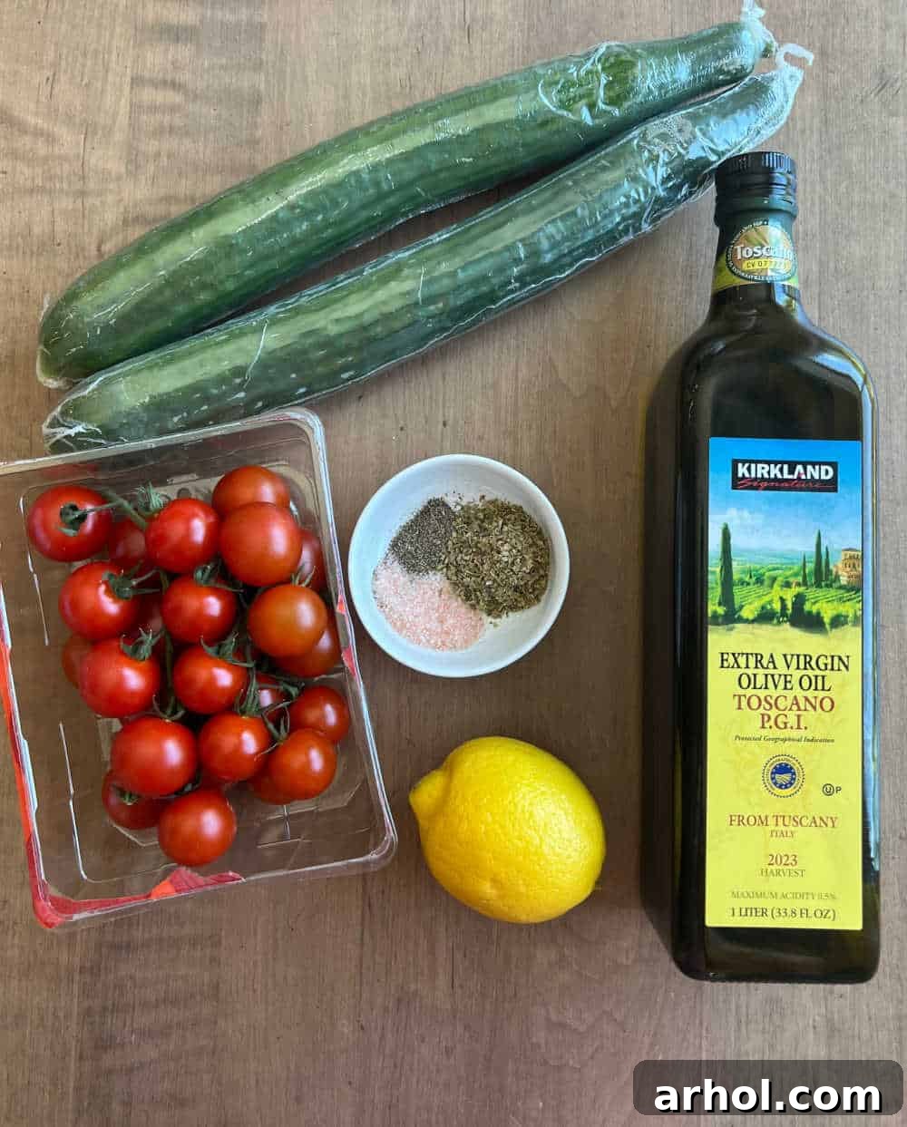 A colorful flat lay of fresh English cucumbers, cherry tomatoes, a whole lemon, a bottle of olive oil, and various seasonings, showcasing the simple ingredients for the cucumber tomato salad.