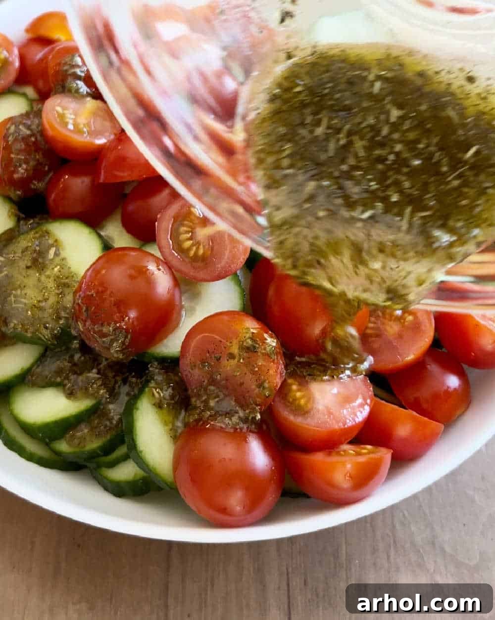 The lemon dressing being poured over a bowl of sliced cucumbers and tomatoes, ready to be mixed and chilled.