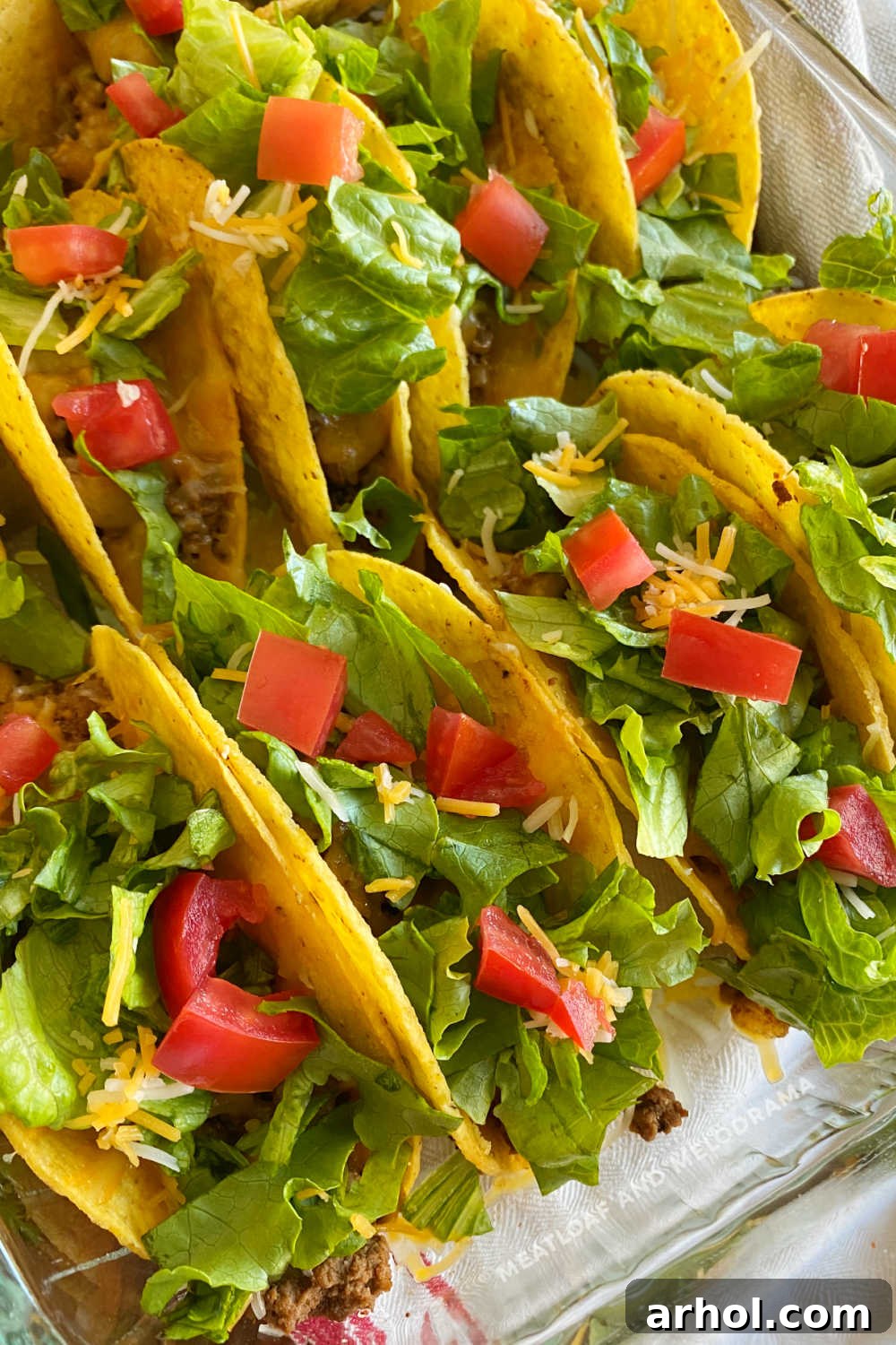 Crispy oven baked beef tacos in a baking dish, garnished with fresh lettuce, diced tomatoes, and a sprinkle of cheese