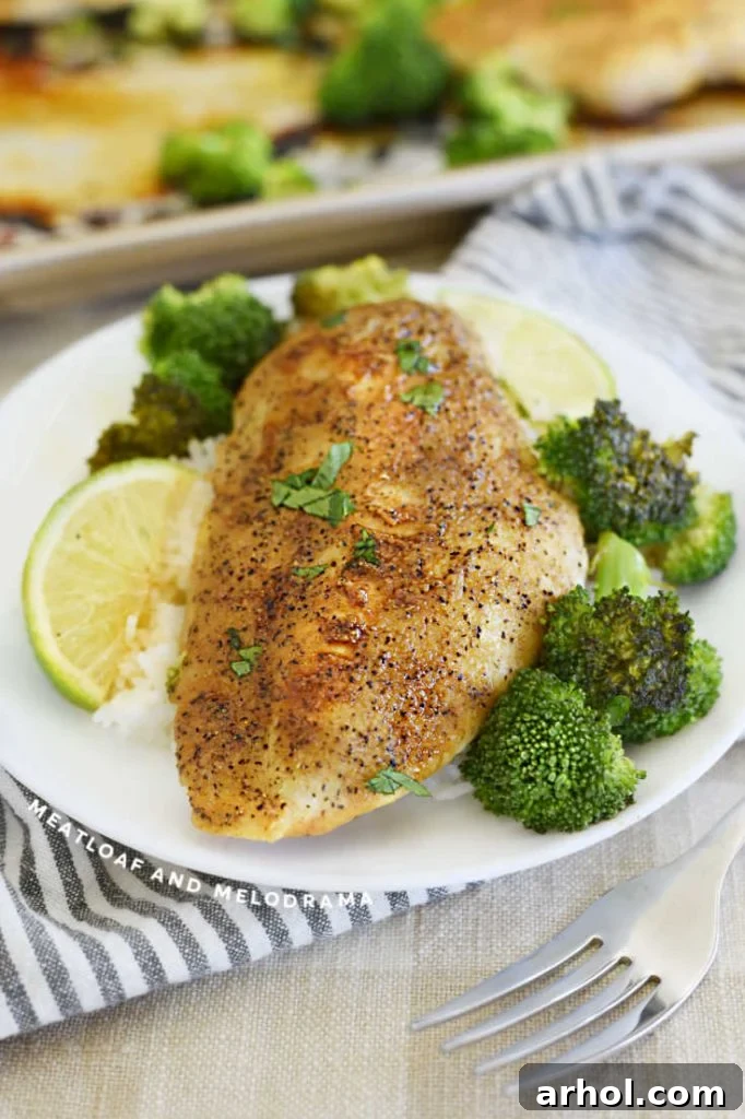 A close-up of a plate with baked honey lime chicken breast, broccoli, and rice, garnished with fresh lime and herbs.
