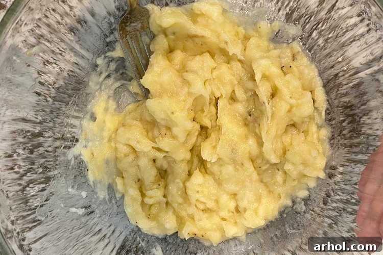 Close-up of three ripe bananas being mashed in a clear glass mixing bowl with a fork, showing their soft, brown-speckled texture.