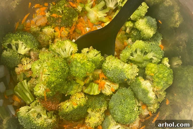 Velvety Instant Pot Broccoli Cheddar Soup 3 A top-down view of fresh broccoli florets and grated carrots mixed with translucent sautéed onions and celery inside the stainless steel pot of an Instant Pot. The vegetables are ready for the addition of broth and subsequent pressure cooking.