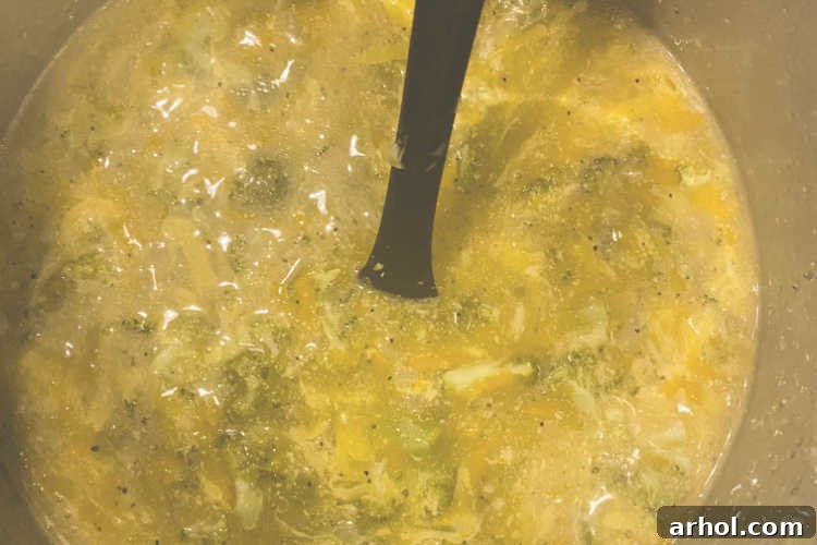 Velvety Instant Pot Broccoli Cheddar Soup 4 A small stream of white cornstarch slurry being poured from a small bowl and stirred into a gently bubbling pot of yellow Instant Pot Broccoli Cheddar Soup with a wooden spoon. The process demonstrates how the soup is thickened to its desired creamy consistency.