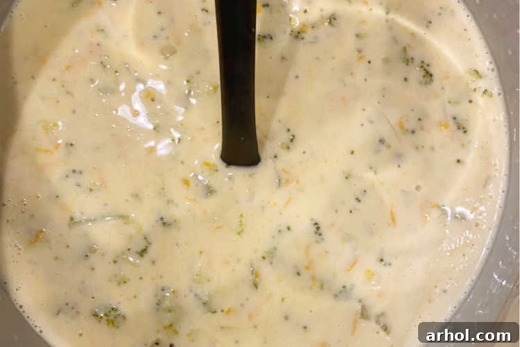 Velvety Instant Pot Broccoli Cheddar Soup 5 A close-up shot of a large wooden spoon stirring heavy cream and a generous amount of freshly grated cheddar cheese into the Instant Pot, which is filled with vibrant yellow Broccoli Cheddar Soup. The cheese is melting, creating a smooth and rich texture.