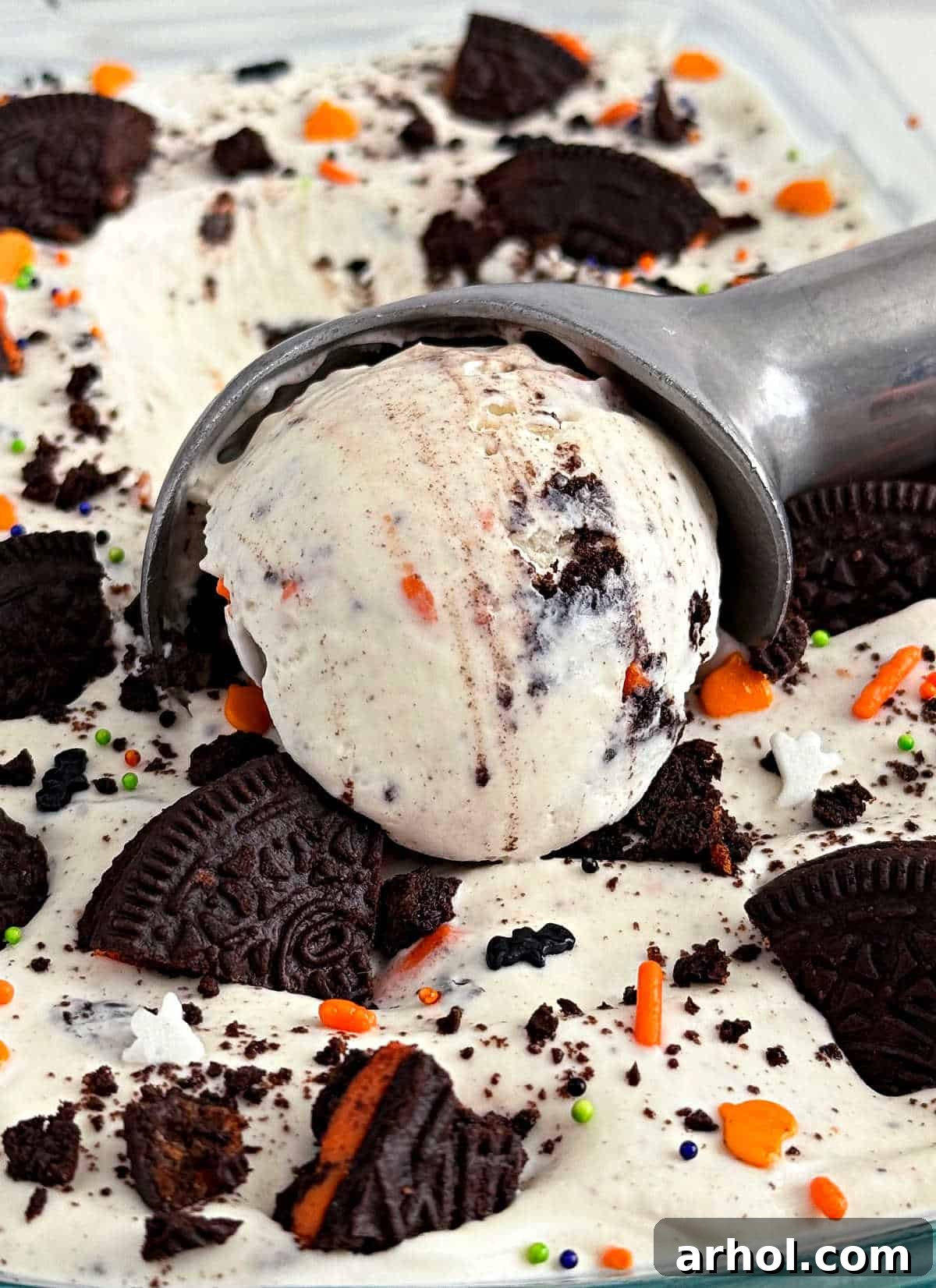 Scooping up vibrant orange and black Halloween Oreo ice cream from a loaf pan, showcasing crushed cookies and sprinkles.