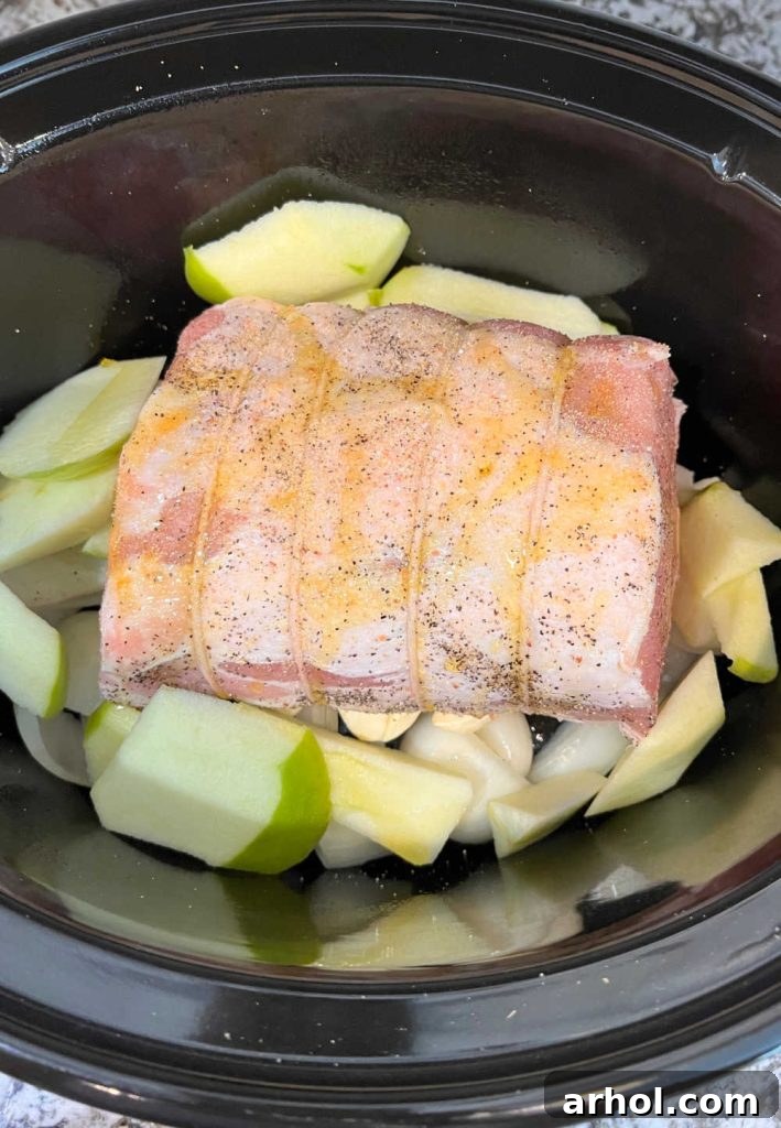 A 4-pound pork loin roast seasoned with Dijon mustard, nestled on a bed of apples and onions in a slow cooker, ready for cooking.