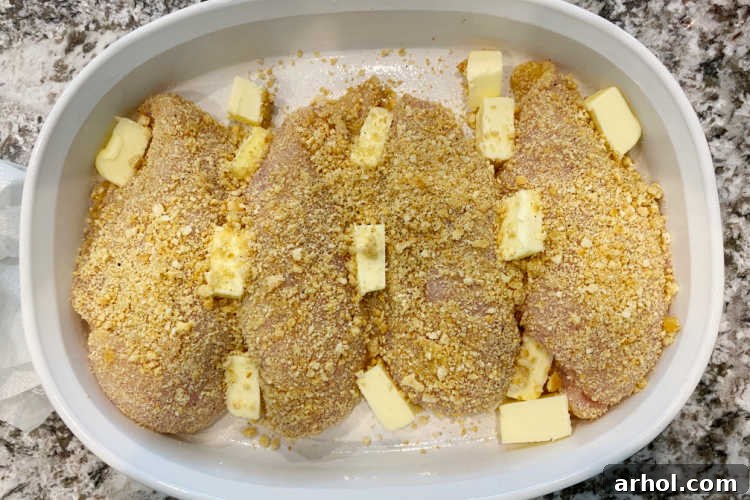 Breaded chicken breasts arranged in a casserole dish with butter cubes placed on top, ready for baking.