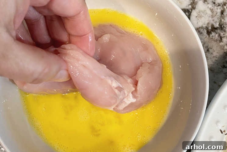 Dipping a raw chicken breast into a bowl of beaten egg wash for coating.