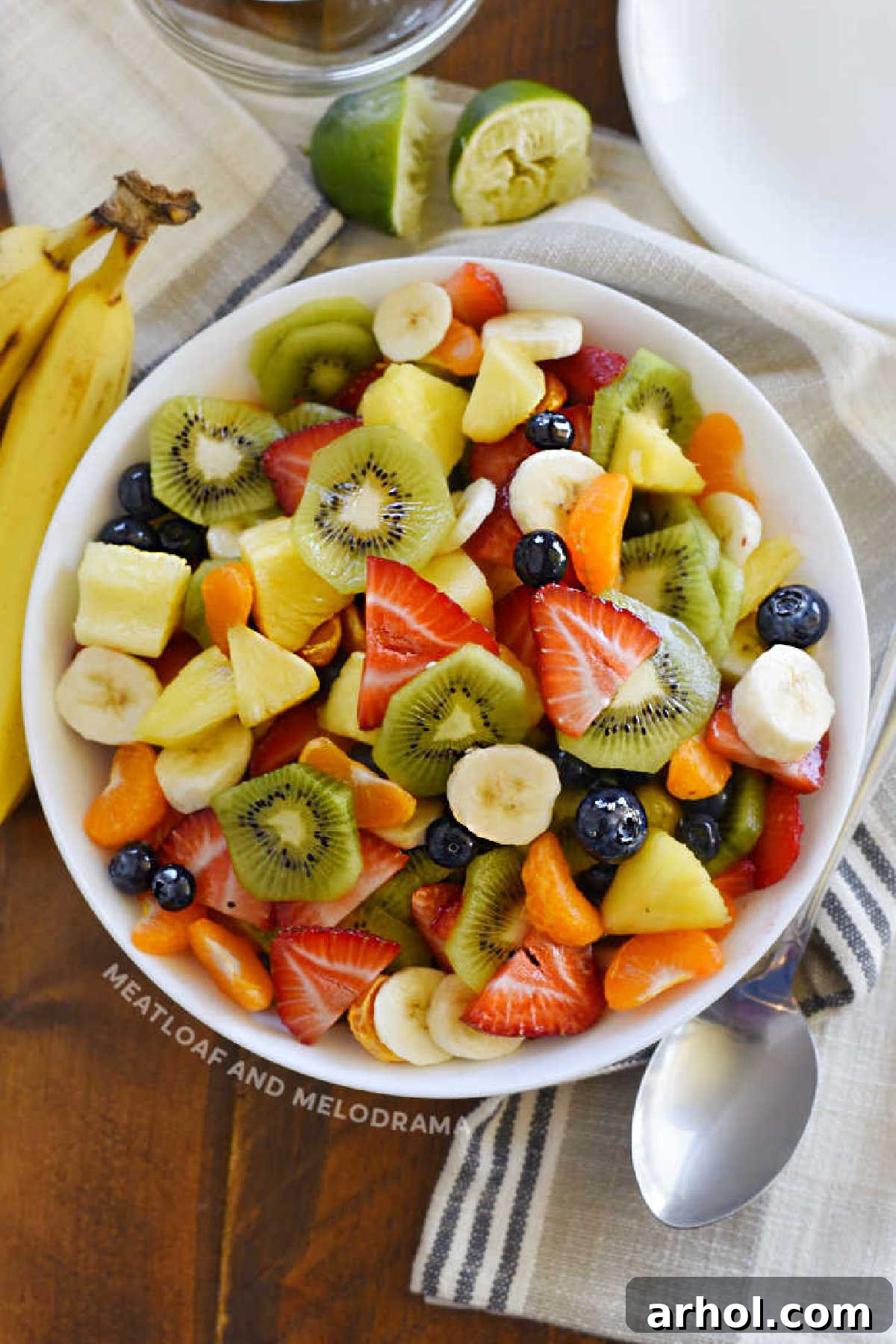 A large bowl brimming with a colorful tropical fruit salad, featuring mandarin oranges, vibrant kiwi slices, ripe bananas, juicy pineapple chunks, fresh strawberries, and sweet blueberries, all glistening with a simple honey lime dressing.